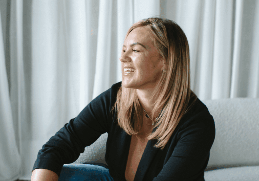 A woman smiling and looking away, sitting casually indoors.
